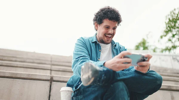 Mosolygós Fiatal Szakállas Férfi Farmeringben Magas Lépcsőn Ülve Mobiltelefont Használva — Stock Fotó