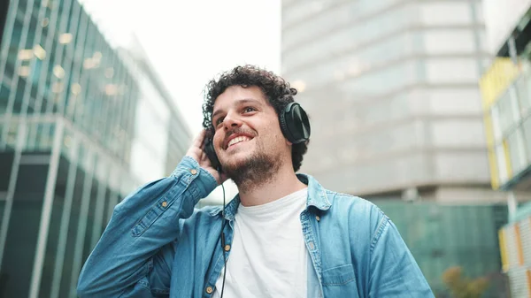 Emotional young bearded man in denim shirt walks down the street in headphones listening to music and sings along, holding mobile phone in his hand on modern cityscape background