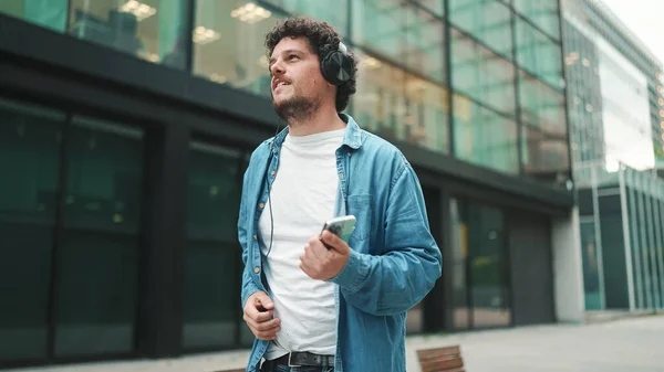 Emotional young bearded man in denim shirt walks down the street in headphones listening to music and sings along, holding mobile phone in his hand on modern cityscape background