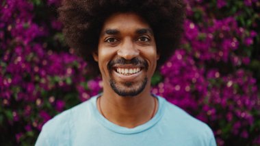 Genç Afro-Amerikalı bir adamın yakın plan portresi gözlerini açar kameraya bakar ve arka plan duvarında mor çiçeklerle kaplı bir gülümsemeyle durur. Kentsel yaşam. 