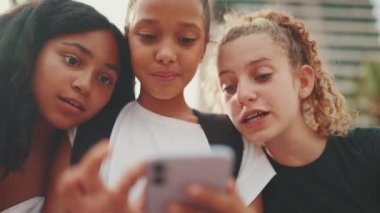 Gençlik öncesi üç kız arkadaş rıhtımda cep telefonu kullanarak oturup video ve fotoğraf izliyorlar. Şehir planında açık havada üç genç var.