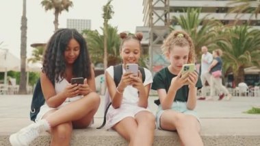 Üç genç kız arkadaş rıhtımda cep telefonuyla oturuyorlar. Gençler videolar, fotoğraflar izliyor, sosyal ağlarda açık hava şehirlerdeki şehirlerin arka planında paylaşıyorlar.