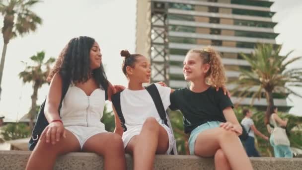 三个十几岁前的女孩朋友坐在海滨笑着 互相拥抱 谈情说爱 城市景观背景下的户外青少年 — 图库视频影像