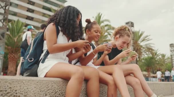三个十几岁前的女孩朋友正坐在海滨用手机 3名青少年在城市景观背景下 在户外用智能手机玩游戏 — 图库视频影像