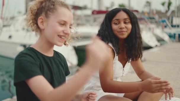 三个青春期前的女朋友坐在海滨 面对着船和游艇的背景 在城市景观的背景下 青少年在户外歌唱和鼓掌 相机在移动 — 图库视频影像