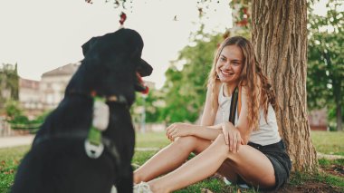 Siyah köpekli tatlı kız çimenlikteki ağacın altında dinleniyor. Mutlu köpek dışarıda yürümekten memnun olur. Açık havada hayvan eğitimi.. 