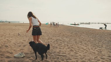 Beyaz bluzlu uzun dalgalı saçlı tatlı kız, çakıl taşı plajında siyah köpek deniz manzaralı oynuyor.