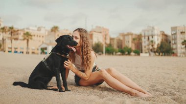 Öğle yemeği vakti. Şirin kız kumsalda evcil hayvanlı sandviçiyle oturuyor. Kız köpeğine sandviçle davranıyor. Modern şehir arka planında sahilde öğle yemeği.