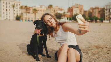 Öğle yemeği vakti. Şirin kız kumsalda evcil hayvanlı sandviçiyle oturuyor. Kız köpeğine sandviçle davranıyor. Modern şehir arka planında sahilde öğle yemeği.