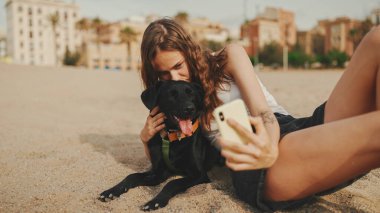 Öğle yemeği vakti. Şirin kız kumsalda evcil hayvanlı sandviçiyle oturuyor. Kız köpeğine sandviçle davranıyor. Modern şehir arka planında sahilde öğle yemeği.