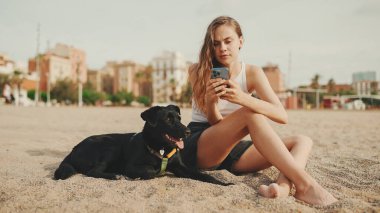 Öğle yemeği vakti. Şirin kız kumsalda evcil hayvanlı sandviçiyle oturuyor. Kız köpeğine sandviçle davranıyor. Modern şehir arka planında sahilde öğle yemeği.