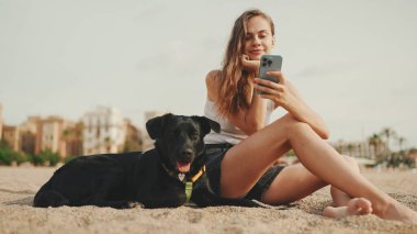 Öğle yemeği vakti. Şirin kız kumsalda evcil hayvanlı sandviçiyle oturuyor. Kız köpeğine sandviçle davranıyor. Modern şehir arka planında sahilde öğle yemeği.