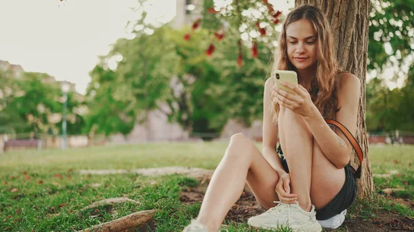 Beautiful girl with long wavy hair wearing in white top uses mobile phone while sitting on the green grass under tree in the park. Girl flip through the pictures on your phone with fingers