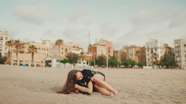 昼食の時間だ 可愛い子はペットのサンドイッチのテストで砂浜に座ってる 少女は犬をサンドイッチで扱う 現代都市の背景にあるビーチでの昼食 — ストック写真