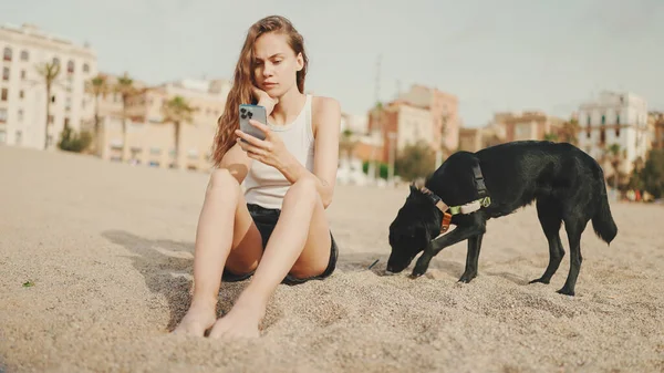 Elinde cep telefonu olan tatlı bir kız modern bina arka planında siyah bir köpekle kumsalda oturuyor. Güzel kız akıllı telefonuyla video ve fotoğraf izliyor.