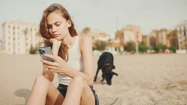 Roztomilá Dívka Mobilním Telefonem Ruce Sedí Písku Pláži Černým Psem — Stock fotografie