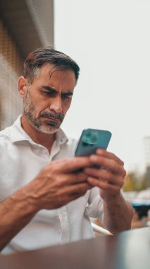 Sakallı, beyaz gömlekli, olgun bir işadamı cep telefonu kullanıyor. Başarılı bir adam kafe masasında oturuyor ve akıllı telefondan yazı yazıyor.