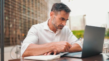 Sakallı olgun bir iş adamı, dizüstü bilgisayarlı bir kullanıcı ve açık kafede oturup notlar alıyor. Doğada açık havada, kafede oturan başarılı bir adam. Mobil ofis serbest.