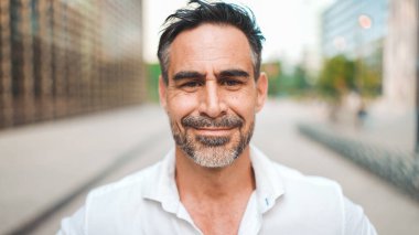 Düzgün sakallı, olgun iş adamı şehrin finans bölgesindeki kameraya bakıyor. Modern binalarda kameraya gülümseyen başarılı bir adam.