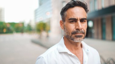Yakın plan iş adamı sakallı, beyaz gömlekli, olgun bir adam şehrin finans bölgesinde başını kameraya çeviriyor.
