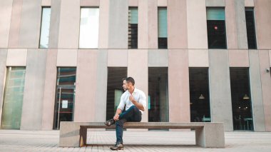 Beyaz gömlek giyen, sakallı, olgun bir iş adamı modern şehrin sokaklarında bankta oturuyor. Başarılı adam dinleniyor.