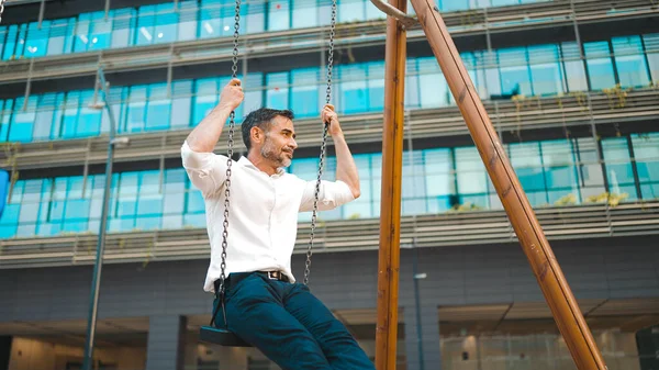 mature businessman with neat beard wearing white shirt swings and rides on children\'s swing. Successful man resting after hard day\'s work