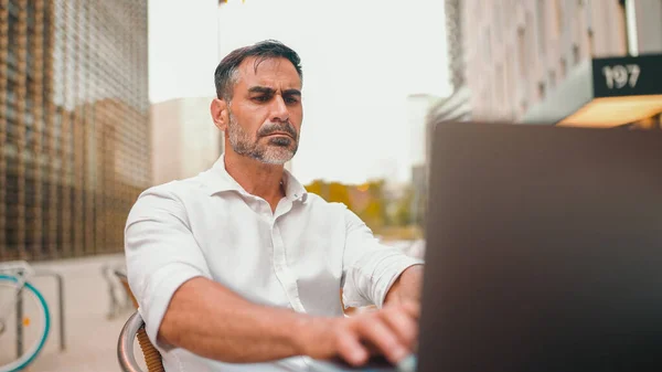 Mature businessman with neat beard wearing white shirt user on laptop pc computer sit at cafe outdoors. Successful man sitting at cafe table outdoors on nature Mobile office freelance