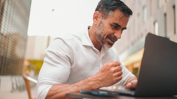 Mature businessman with neat beard wearing white shirt user on laptop pc computer sit at cafe outdoors. Successful man sitting at cafe table outdoors on nature Mobile office freelance