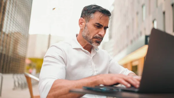 Mature businessman with neat beard wearing white shirt user on laptop pc computer sit at cafe outdoors. Successful man sitting at cafe table outdoors on nature Mobile office freelance