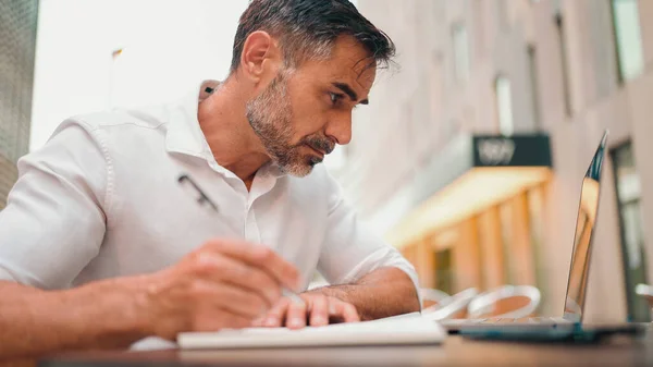 Mature businessman with neat beard wearing white shirt writing down notes in paper notepad, sitting at table. making appointments daily tasks in personal organizer