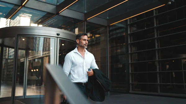 Mature businessman with neat beard wearing white shirt leaves the office in the financial district in the city. Successful man with mobile phone in his hands, after hard day\'s work