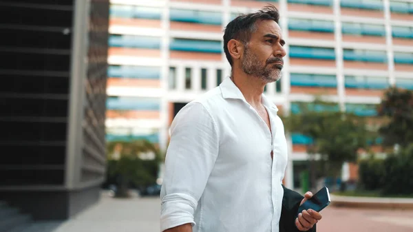 Mature businessman with neat beard wearing white shirt leaves the office in the financial district in the city. Successful man with mobile phone in his hands, after hard day\'s work