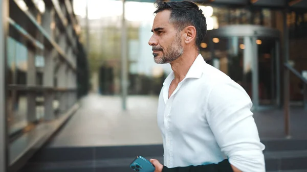 Mature businessman with neat beard wearing white shirt leaves the office in the financial district in the city. Successful man with mobile phone in his hands, after hard day\'s work