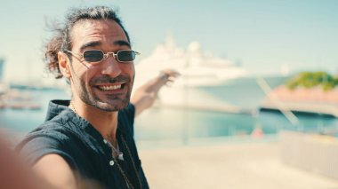Uzun kıvırcık saçlı ve sakallı genç İtalyan cep telefonuyla selfie çekiyor. Güneş gözlüklü şık bir adam akıllı telefonunun kamerasına gülümsüyor ve limana demirlemiş yatı işaret ediyor.