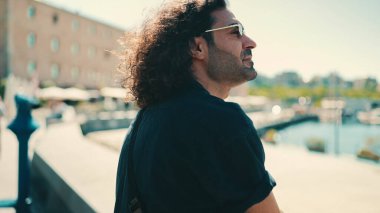 Uzun kıvırcık saçlı ve sakallı genç İtalyan limanda oturur ve gemilere ve yatlara bakar.