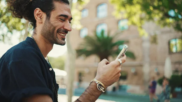 Attractive italian guy with ponytail and stubble is using mobile phone sitting on bench at old buildings background. Stylish man writes message, views photo on smartphone