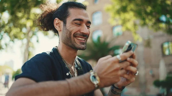 Attractive italian guy with ponytail and stubble is using mobile phone sitting on bench at old buildings background. Stylish man writes message, views photo on smartphone