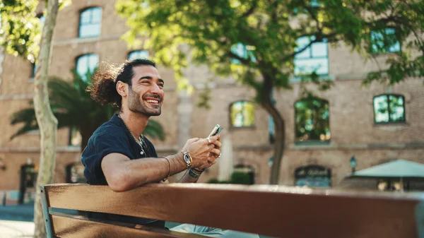 Attractive italian guy with ponytail and stubble is using mobile phone sitting on bench at old buildings background. Stylish man writes message, views photo on smartphone