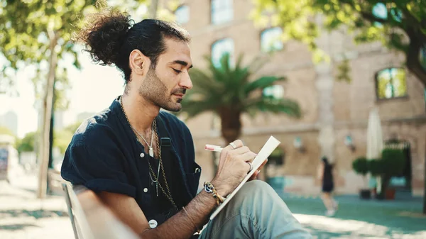 Young italian guy with ponytail and stubble sits on street bench and makes sketches with pen on piece of paper