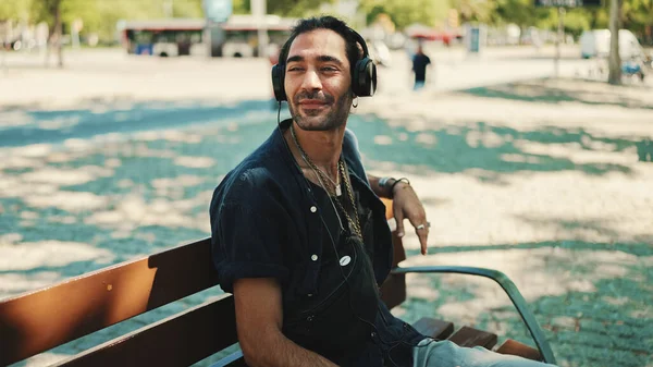 Closeup of young italian guy with ponytail and stubble sits with headphones on street bench and listens to music on cityscape background