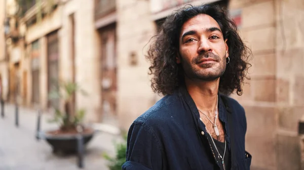 Young italian guy with long curly hair and stubble looks around looking at the architecture of the city. stylish man with an earring in his ear and lot of chains. Tourist looking at the sights