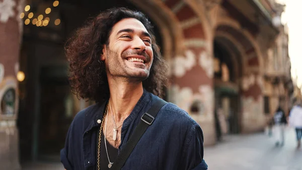 Young italian guy with long curly hair and stubble looks around looking at the architecture of the city. stylish man with an earring in his ear and lot of chains. Tourist looking at the sights