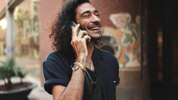 Young attractive italian guy with long curly hair and stubble is using mobile phone at old buildings background. Stylish man with an earring in his ear and lot of chains emotionally talking on phone