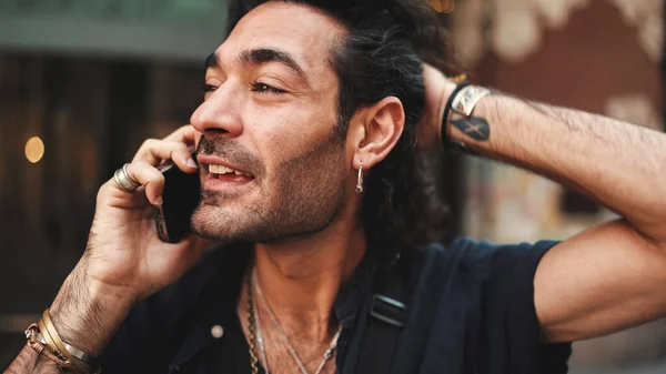 Young attractive italian guy with long curly hair and stubble is using mobile phone at old buildings background. Stylish man with an earring in his ear and lot of chains emotionally talking on phone
