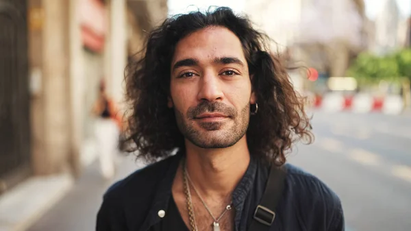 Young attractive italian guy with long curly hair and stubble looks into the camera and smiles at old buildings background. Stylish man with an earring in his ear and lot of chains