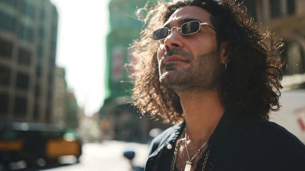 Young italian guy with long curly hair and stubble walks along the fenced sidewalk and looks around.