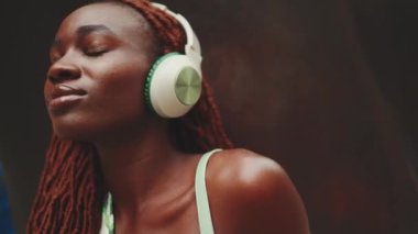 Kulaklıklı Afrikalı güzel bir kadının sokakta müzik dinleyerek eğlendiği yakın plan çekimler. Şık kız gözleri kapalı müzik dinliyor.
