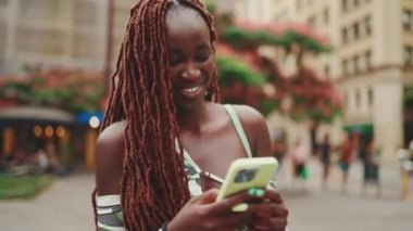 Üzerinde Afrika örgüleri olan gülümseyen muhteşem bir kadın sokakta dikiliyor ve cep telefonu kullanıyor. Şık kız akıllı telefondan mesaj yazıyor.