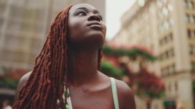 Giyinip süslenmiş, Afrika örgülü muhteşem bir kadın caddede yürüyor. Havalı gülümseyen kız binaların üst katlarına bakıyor. Kamera yakınlaştırması