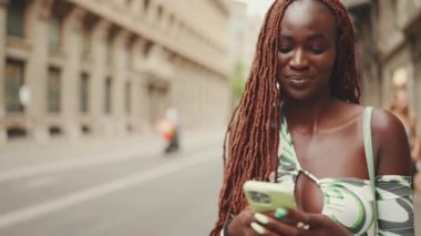 Sırıtkan, Afrika örgülü muhteşem bir kadın kaldırımda duruyor ve cep telefonu kullanıyor. Şık kız akıllı telefondan mesaj yazıyor.